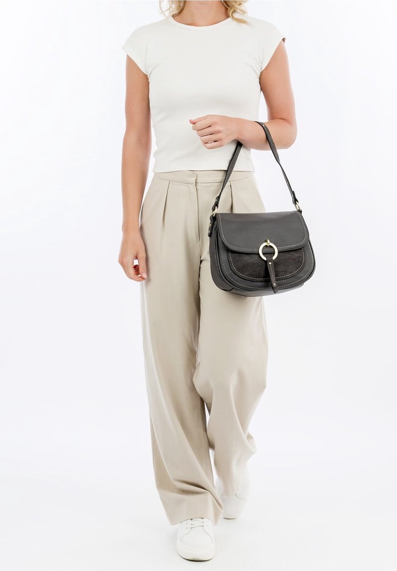 Femme portant un t-shirt blanc et un pantalon large beige, portant un sac à épaule en cuir noir avec détail anneau en métal.