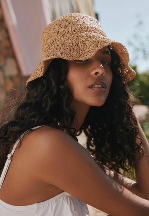 Woman with curly black hair wearing a tan crocheted bucket hat and white sleeveless top, looking toward the camera outdoors in sunlight.