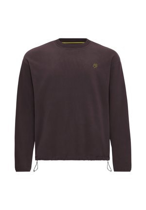 Giovane uomo con capelli ricci che indossa una felpa marrone scuro a maniche lunghe e pantaloni neri, rivolto leggermente di lato.