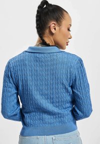 Blå strikket cardigan med et teksturert mønster, kragehals, ribbestrikket kant og elastiske mansjetter. Bakvinduet viser design detaljer.