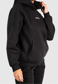 Personne portant un sweat à capuche noir avec une poche frontale et un petit texte blanc sur la poitrine, tenant le col du sweat à capuche d'une main.