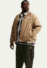 Homme debout portant une veste Nike beige, une chemise à carreaux noir et blanc, un sous-vêtement blanc et un pantalon Nike noir, regardant sur le côté.