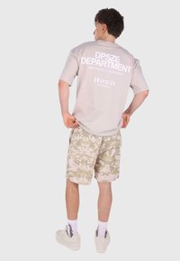 Hellbeige Oversize-T-Shirt mit großen weißen Druck auf dem Rücken, kombiniert mit beigen Camouflage Shorts und weißen Sneakers.
