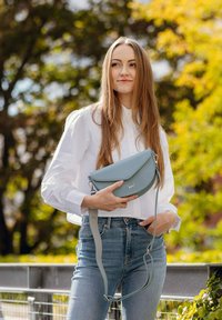 Měkká modrá kožená kabelka přes rameno s přeloženou klopou, stříbrným kováním a odnímatelným popruhem, držená osobou v bílé blůze.