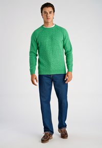 Suéter verde de punto con patrones trenzados, cuello redondo y puños acanalados. Combinado con jeans azules y zapatos náuticos marrones. Look sencillo y casual.