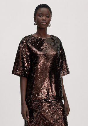 Woman wearing a shiny, dark brown sequin top and matching skirt, with short natural hair and silver hoop earrings, standing against plain background.