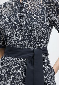 Φόρεμα ναυτικού με λευκά κεντημένα αφηρημένα μοτίβα φύλλων, κουμπιά καλυμμένα με ύφασμα στο μπροστινό μέρος και φαρδιά ζώνη στο ίδιο χρώμα δεμένη στη μέση.