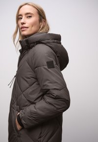 Doudoune grise avec capuche, présentant un design matelassé, des poignets élastiques, des poches latérales et un patch de marque noir sur la manche gauche.