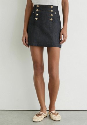 Kvinde iført mørk denim mininederdel med seks dekorative guldknapper kombineret med beige vævede flade sko mod en ensfarvet baggrund.