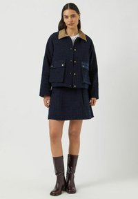 Mörkblå jacka med en tan-färgad krage, två framfickor och dekorativ sömnad, kombinerad med en matchande mörkblå kjol och glänsande bruna stövlar.
