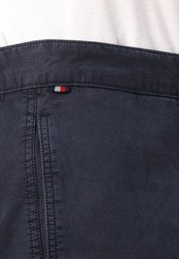 Kobaltblauwe stof met een gladde textuur, voorzien van een klein rood, wit en blauw label op de achterkant, boven een verticale naad. Minimalistisch ontwerp.