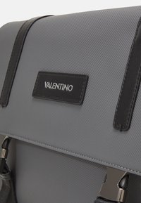 Šedá texturovaná taška s černými koženými akcenty. Obsahuje lesklou destičku s logem "Valentino", nastavitelné ramenní popruhy a kovové příslušenství.