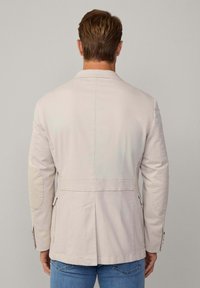 Beige Jacke mit strukturiertem Design, ausgestattet mit Ellbogenpatches, einem einzelnen Rückenschlitz und zwei seitlichen Taschen. Aus glattem Stoff gefertigt.