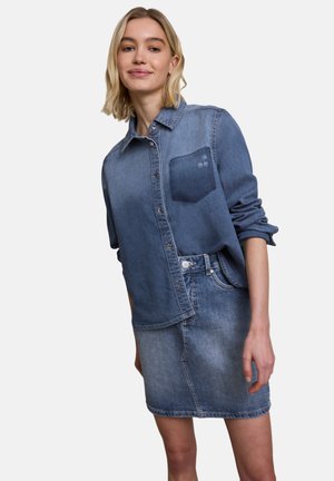 Blonde Frau trägt ein blaues Jeanshemd mit aufgekrempelten Ärmeln und einen ausgebleichten blauen Jeansrock, steht vor einem einfarbigen Hintergrund.