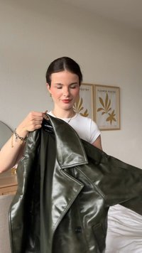 Kurtka ze skóry w oliwkowej zieleni, z dwurzędowym zapięciem, klapami z wycięciem i błyszczącą fakturą, trzymana przez osobę w jasnej koszuli.