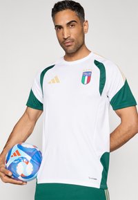 Biely futbalový dres so zelenými prvkami, logom Adidas, erbom talianskej futbalovej reprezentácie, krátkymi rukávmi, textúrovanou látkou a zlatými pruhmi na ramenách.