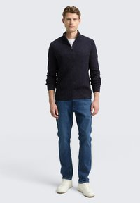 Mann trägt einen marineblauen Pullover mit Viertelreißverschluss, blaue Jeans und weiße Sneaker, steht vor einem einfarbigen hellen Hintergrund.