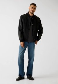 Veste bomber noire avec manches en cuir, fermeture à pression et poignets côtelés, associée à un jean bleu clair et des chaussures noires.