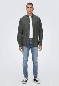 Homem de pé, vestindo uma camisa cinza escura com botões sobre uma t-shirt branca, jeans rasgados de um azul claro e ténis pretos com atacadores brancos.