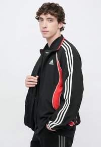 Čierna bunda s červenými akcentmi, bielymi troma pruhmi na rukávoch, predným zipsom a elastickými manžetami. Materiál pôsobí hladko a ľahko.