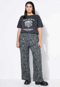 Zwarte graphic t-shirt met bloemmotief gecombineerd met paarse en groene wijde broek met bloemenpatroon. Model draagt chunky zwarte schoenen.