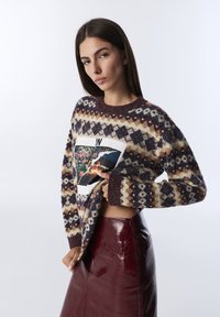 Woman wearing a patterned sweater with a floral and fire graphic and a burgundy leather skirt, posing against a plain light background.