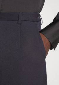 Pantalon bleu marine à texture lisse, avec passants de ceinture, poches avant et coupe ajustée ; assorti à un poignet de chemise noir.