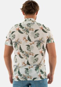 Man met een korte mouwen bloemenoverhemd met groene bladeren en oranje bloemen, gezien vanachter, gecombineerd met blauwe jeans.