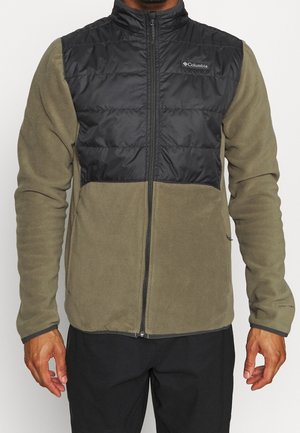 Mann trägt eine Jacke mit Reißverschluss mit olivgrünen Fleeceärmeln und -körper sowie einem schwarzen, gesteppte Brustpaneel mit Columbia-Logo.