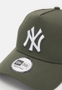 Casquette vert olive en tissu avec un logo "NY" brodé en blanc, une sangle réglable et un label circulaire "New Era" sur le bord.