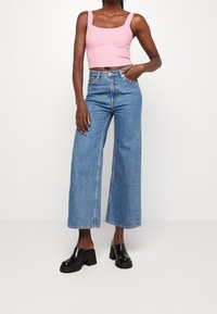 Lichtroze cropped tanktop met vierkante hals, gecombineerd met hoge taille en wijde blauwe jeans. Zwarte platform mules accentueren de outfit.