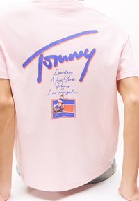 Camiseta de color rosa claro con el logotipo "Tommy" en azul y naranja, nombres de ciudades como Londres, Nueva York, París, Los Ángeles y un pequeño gráfico de un velero en la parte de atrás.