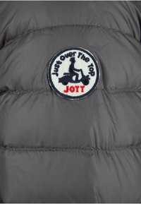 Chaqueta acolchada gris con un parche redondo bordado que presenta un diseño de scooter y el texto "Just Over the Top JOTT" en negro y rojo.