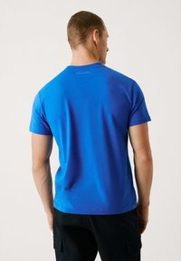 Sinine puuvillane T-Särk lühikeste варukatega, ümmarguse kaelusega ja "Pepe Jeans" logo trükiga seljal, koos mustade pükstega.