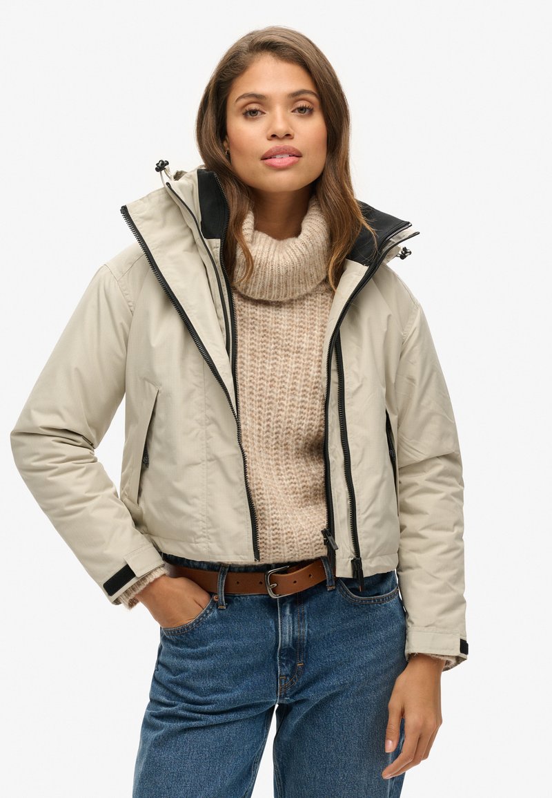 Beżowa krótka kurtka z czarnymi akcentami, z zamkami z przodu i kapturem, noszona na kremowym swetrze z golfem, w połączeniu z niebieskimi dżinsami i brązowym paskiem.