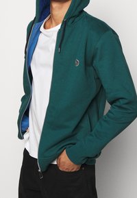 Türkise Zip-Hoodie mit blauer Innenseite, elastischen Bündchen und Kordelzügen. Verfügt über ein kleines Logo auf der Brust. Getragen über einem weißen T-Shirt.