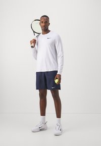 Camisa blanca de manga larga con botones, pantalones cortos azul marino con logo, sosteniendo una raqueta de tenis y una pelota, vistiendo zapatillas deportivas blancas y calcetines.