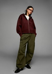 Woman wearing a burgundy zip-up hoodie, white shirt, olive green cargo pants, black loafers, and glasses, standing against a gray background.