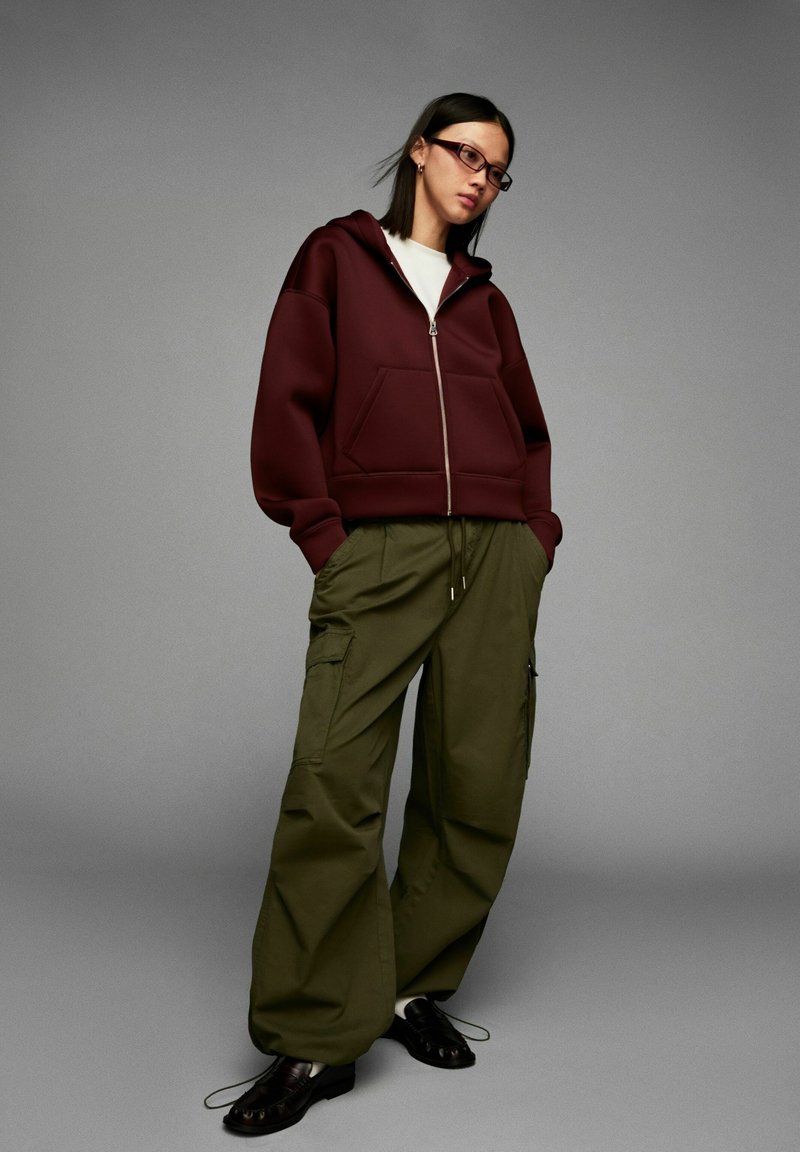 Woman wearing a burgundy zip-up hoodie, white shirt, olive green cargo pants, black loafers, and glasses, standing against a gray background.