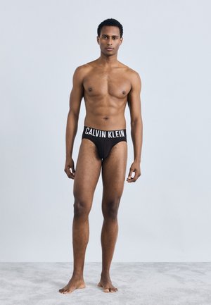 Μαύρα σλιπ Calvin Klein με ελαστική ζώνη που έχει αντίθετες λευκές επιγραφές. Το μοντέλο έχει λεπτή σιλουέτα και στέκεται ξυπόλητο σε μια ανοιχτό γκρίζα επιφάνεια.