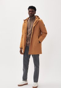 Parka impermeable para hombre en tono tostado, con capucha, cierre de cremallera y bolsillos laterales, utilizado sobre un suéter beige de cuello con cremallera, combinado con jeans oscuros y zapatillas blancas.