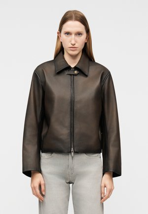 Γυναίκα με ίσια ανοιχτό καφέ μαλλιά φοράει καφέ σκούρο δερμάτινο μπουφάν με φερμουάρ μπροστά και ανοιχτό γκρι τζιν, στέκεται μπροστά σε απλό φόντο.