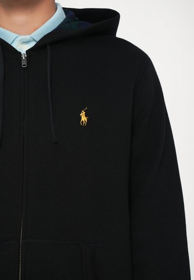 Sweatshirt preto com fecho éclair, feito de tecido macio, apresentando um logo bordado a ouro no lado esquerdo do peito e um forro em padrão de xadrez azul na capuz.