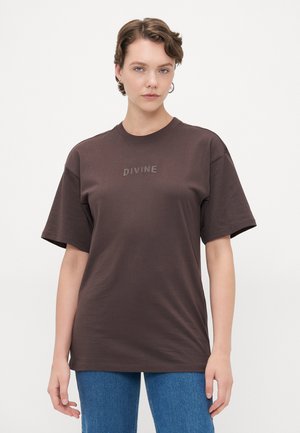 Brun bomull T-skjorte med korte ermer, rund hals, og teksten "DIVINE" i en lysere nyanse på forsiden. Brukt med blå jeans.