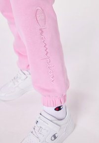 Rozā sporta bikses ar elastīgām aprocēm, ar izšūtu "Champion" logotipu. Apvienotas ar baltām sporta apaviem un baltiem zeķēm ar logotipu.