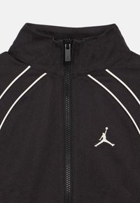 Μαύρο αθλητικό μπουφάν με σταθερό γιακά, πλήρη φερμουάρ, άσπρες ραφές και εξέχον λευκό λογότυπο Jumpman στην αριστερή πλευρά. Λείος υφασμάτινος σχεδιασμός.