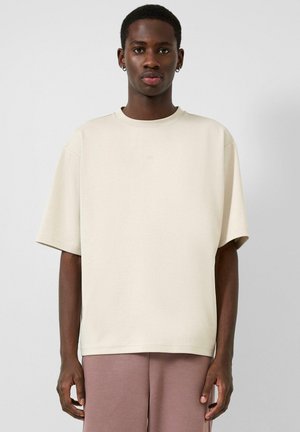 Homme à la peau foncée portant un T-shirt beige surdimensionné et un pantalon mauve, debout devant un fond uni gris clair.