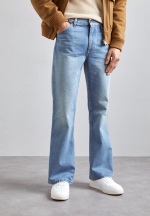 Mann trägt hellblaue Schlagjeans, weiße Turnschuhe, beigen Pullover und braune Jacke und steht auf grauem Boden vor einer schlichten Wand.