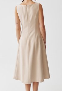 En ärmlös klänning i knä längd av beige tyg med en slät textur, har en blygsam halsringning och en mitt bak dragkedja.