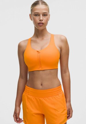 Femme portant un soutien-gorge de sport zippé orange vif et un short de sport orange assorti sur un fond uni.